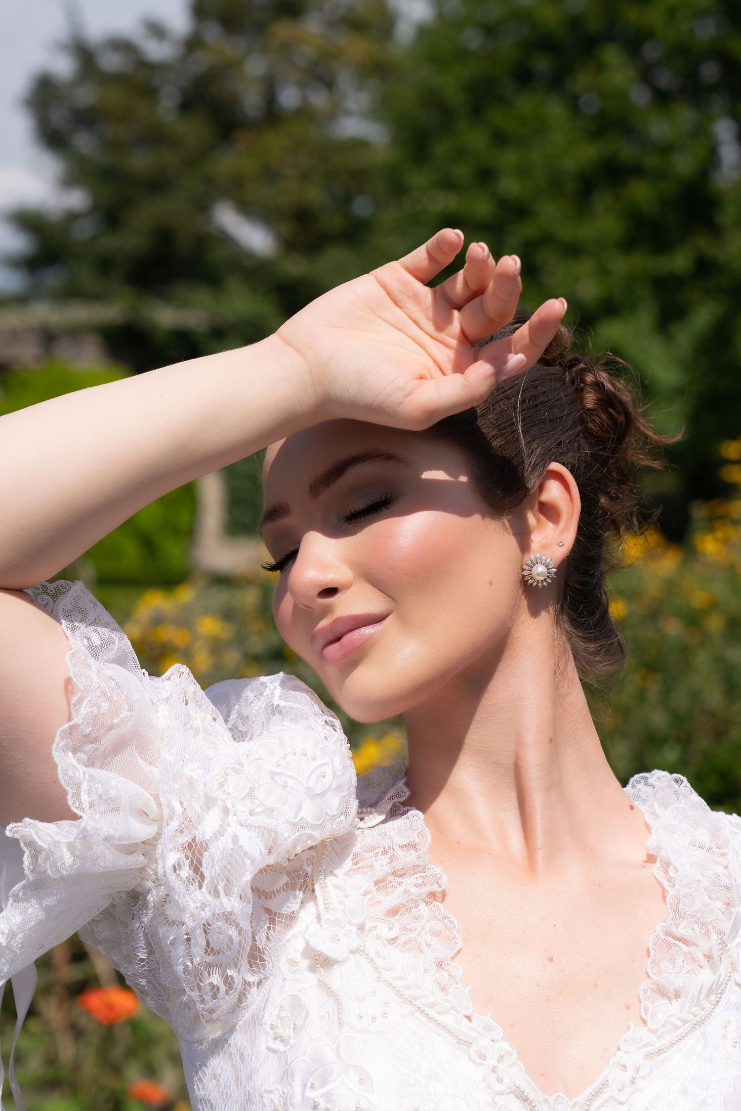 Beautiful bride in lace wedding dress shields her eyes from bright sunlight in a colorful garden setting.