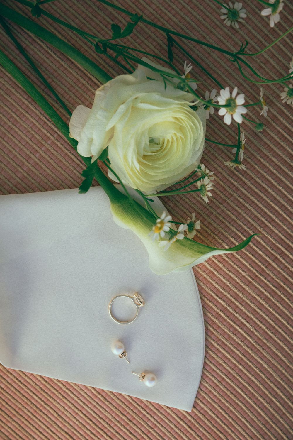 Wedding flat lay featuring a white ranunculus, pearl earrings, and a gold ring on a white envelope.