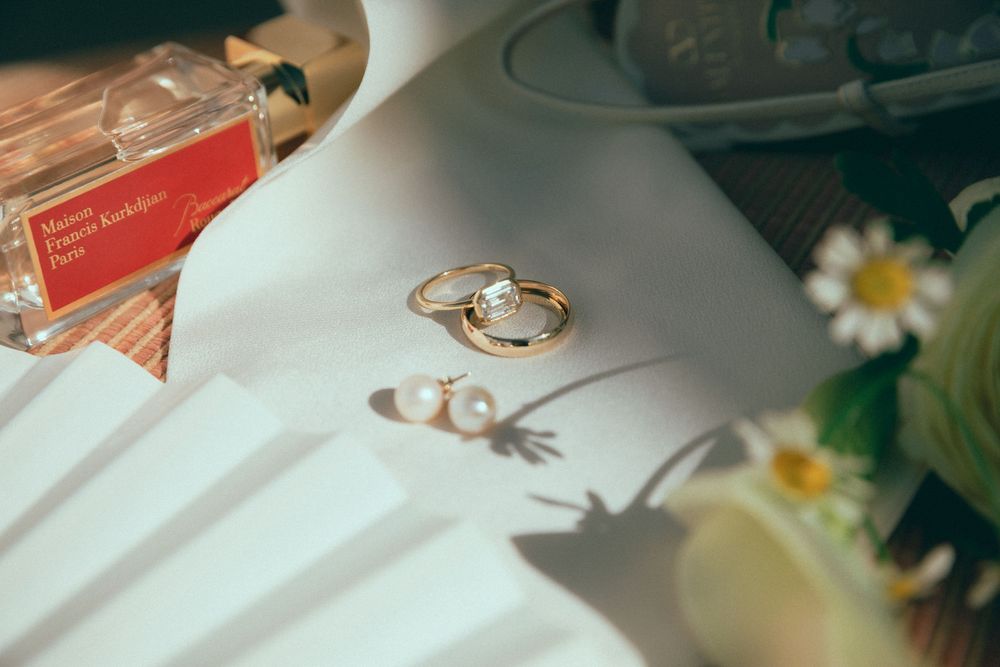 Gold rings and pearl earrings on white surface beside Maison Kitsune perfume bottle and daisy flowers.
