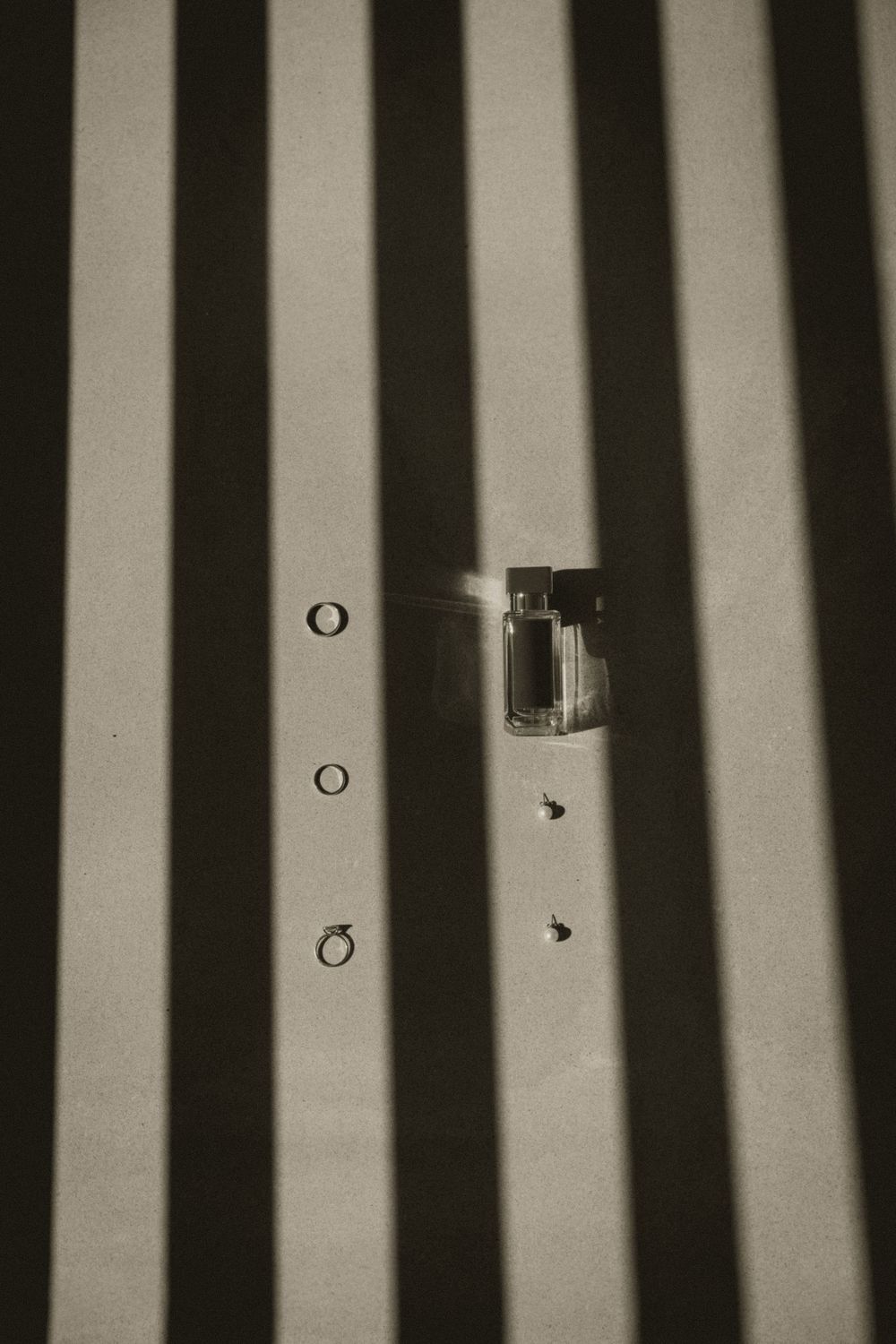 Black and white photo of wedding rings and perfume bottle with dramatic striped shadows on a flat surface.
