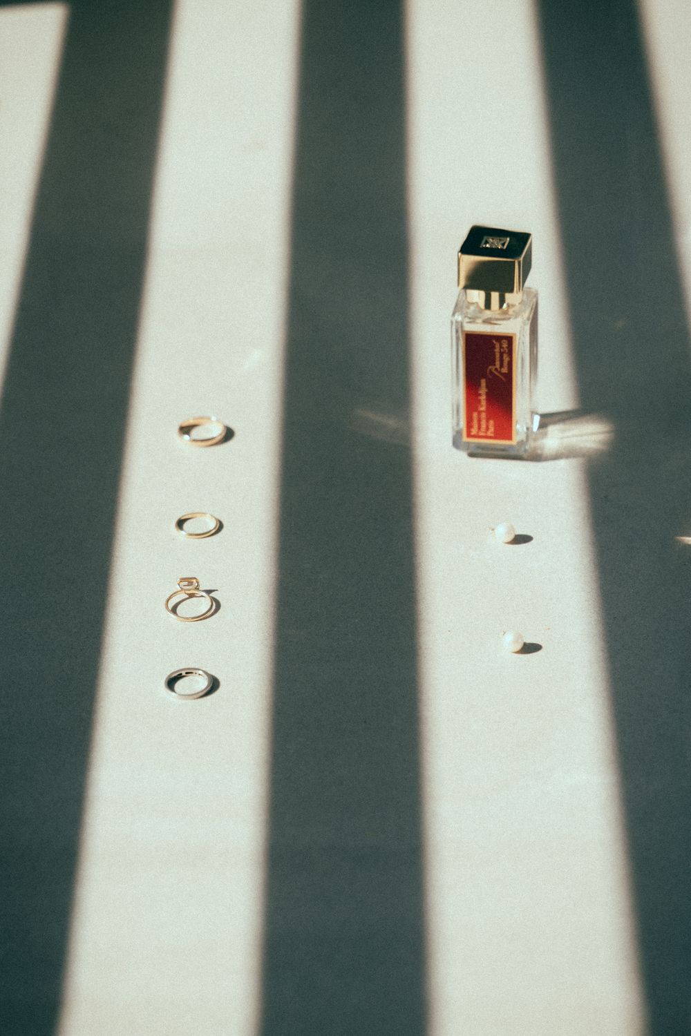 Wedding rings and a Maison Francis Kurkdjian perfume bottle arranged on a striped surface with dramatic shadow play.