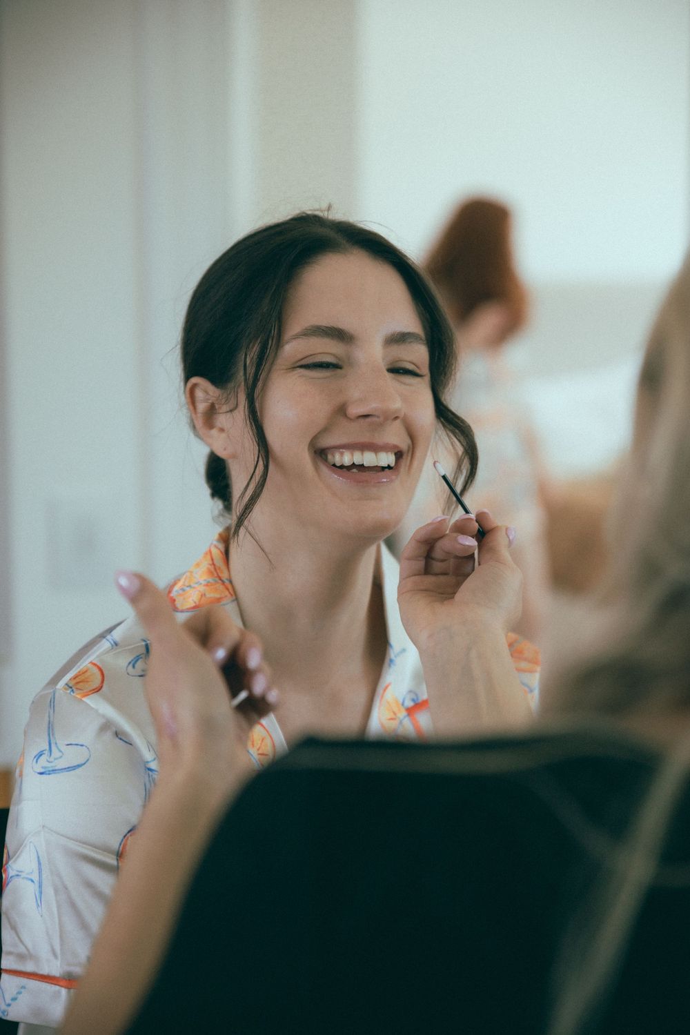Bride laughing joyfully while getting makeup applied during wedding morning preparations.
