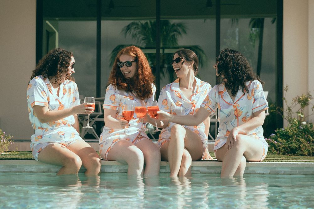 Four women in matching pajamas sit poolside, dangling feet in water while toasting with pink drinks on a sunny day.