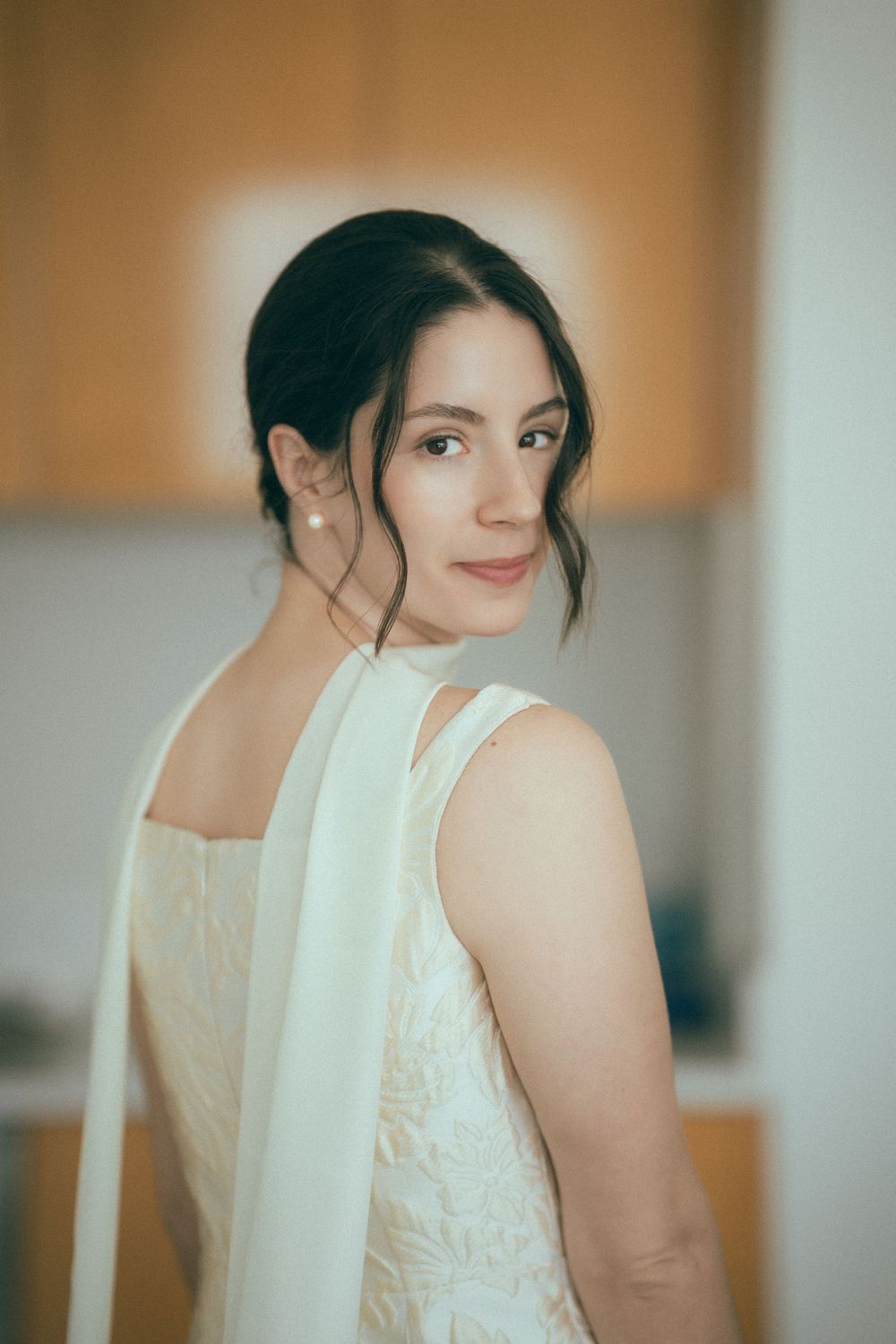 A young woman in a white lace dress with a flowing sash looks over her shoulder, wearing pearl earrings.