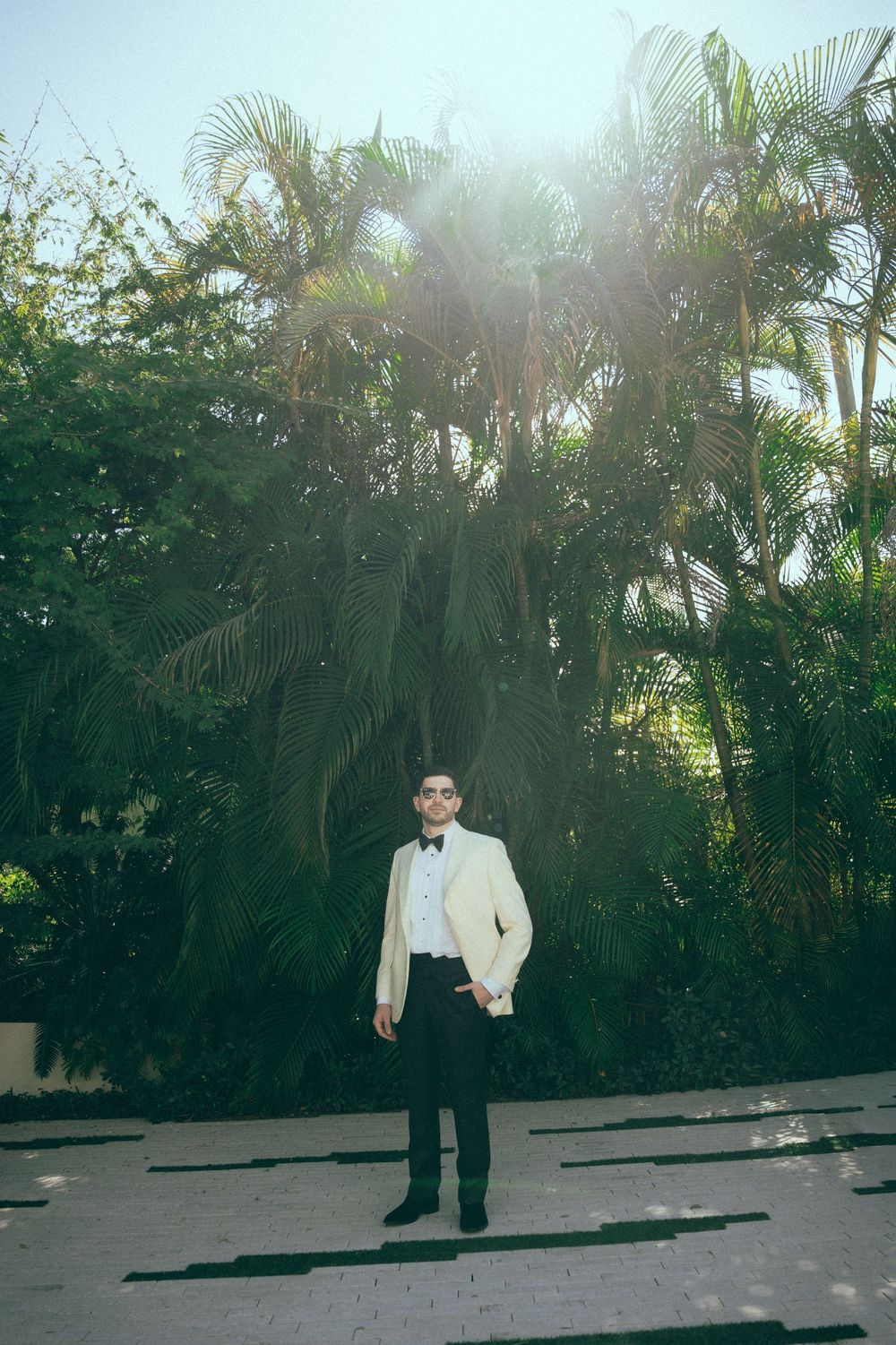 Groom in white tuxedo jacket standing outdoors among tropical palm trees in sunlight.