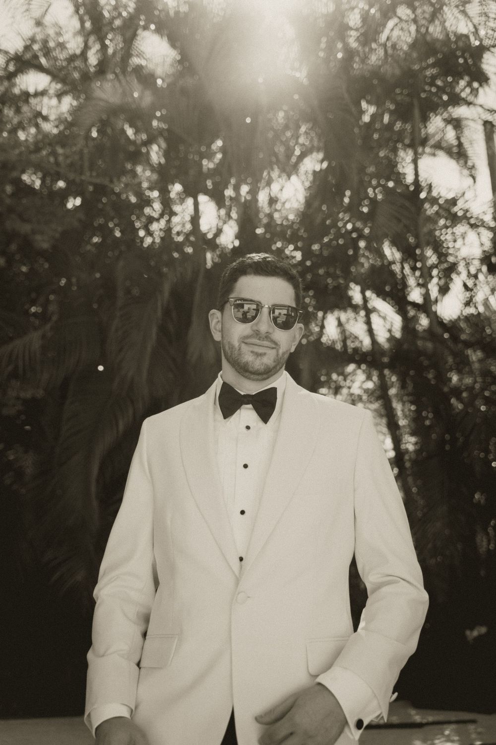 A groom in a white tuxedo and black bow tie poses outdoors in a sepia-toned photo with sunglasses on.