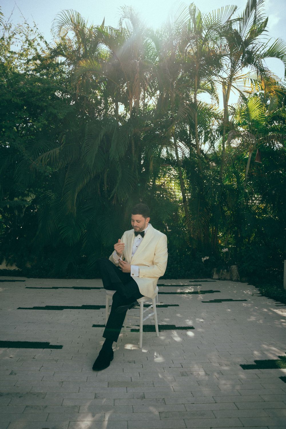 Groom in white tuxedo jacket and bow tie sits on chair outdoors surrounded by lush tropical palm trees.