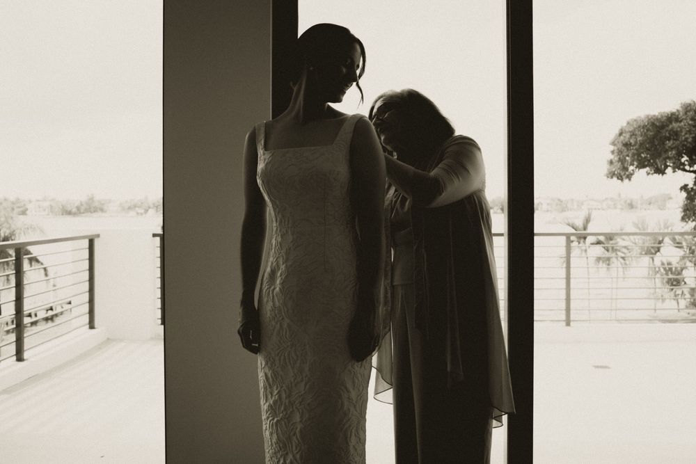 Black and white photo of bride in lace wedding dress embracing an emotional guest by a balcony window.