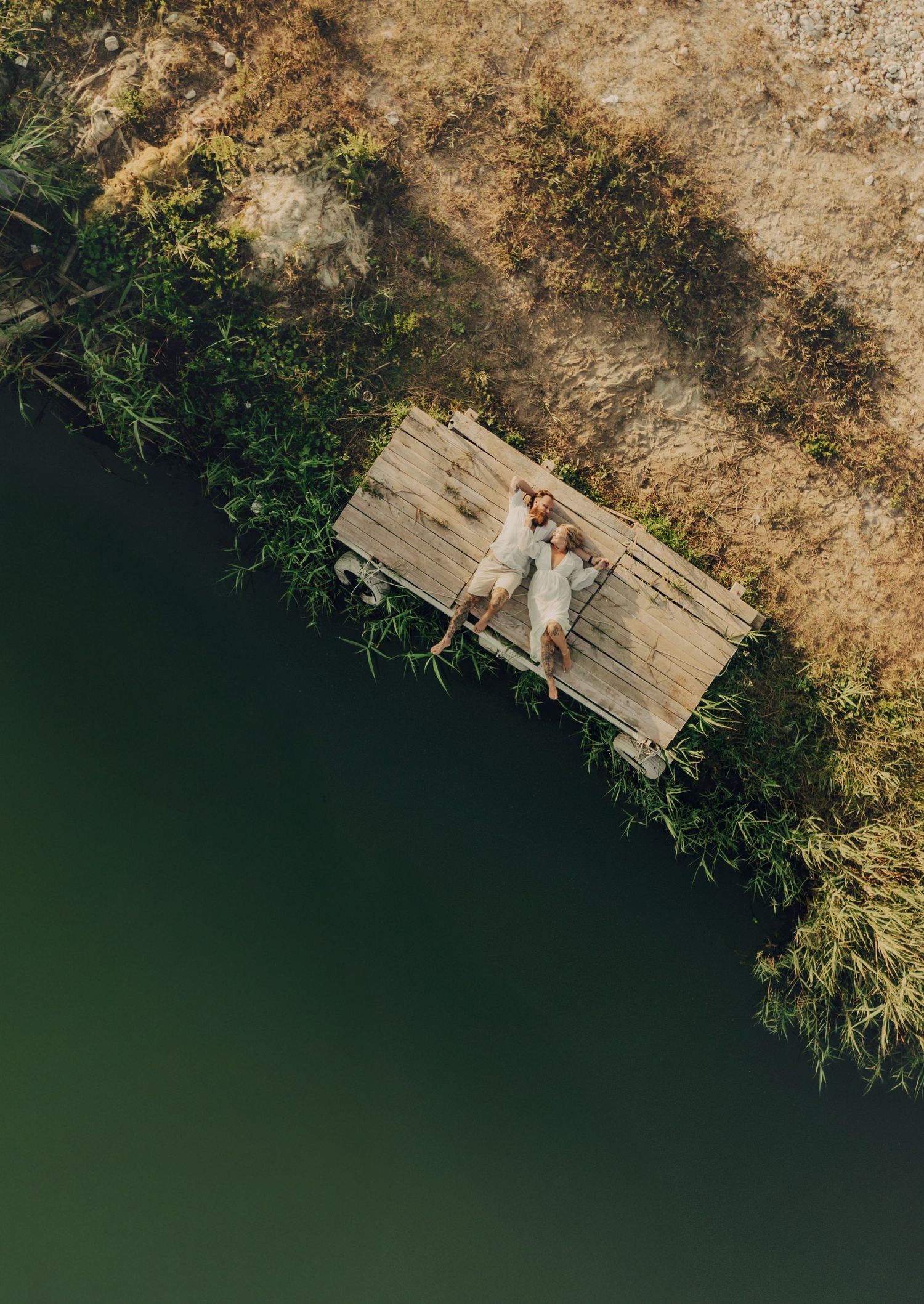 Aerial drone view of a couple lying on a wooden dock beside a calm green lake surrounded by lush vegetation.