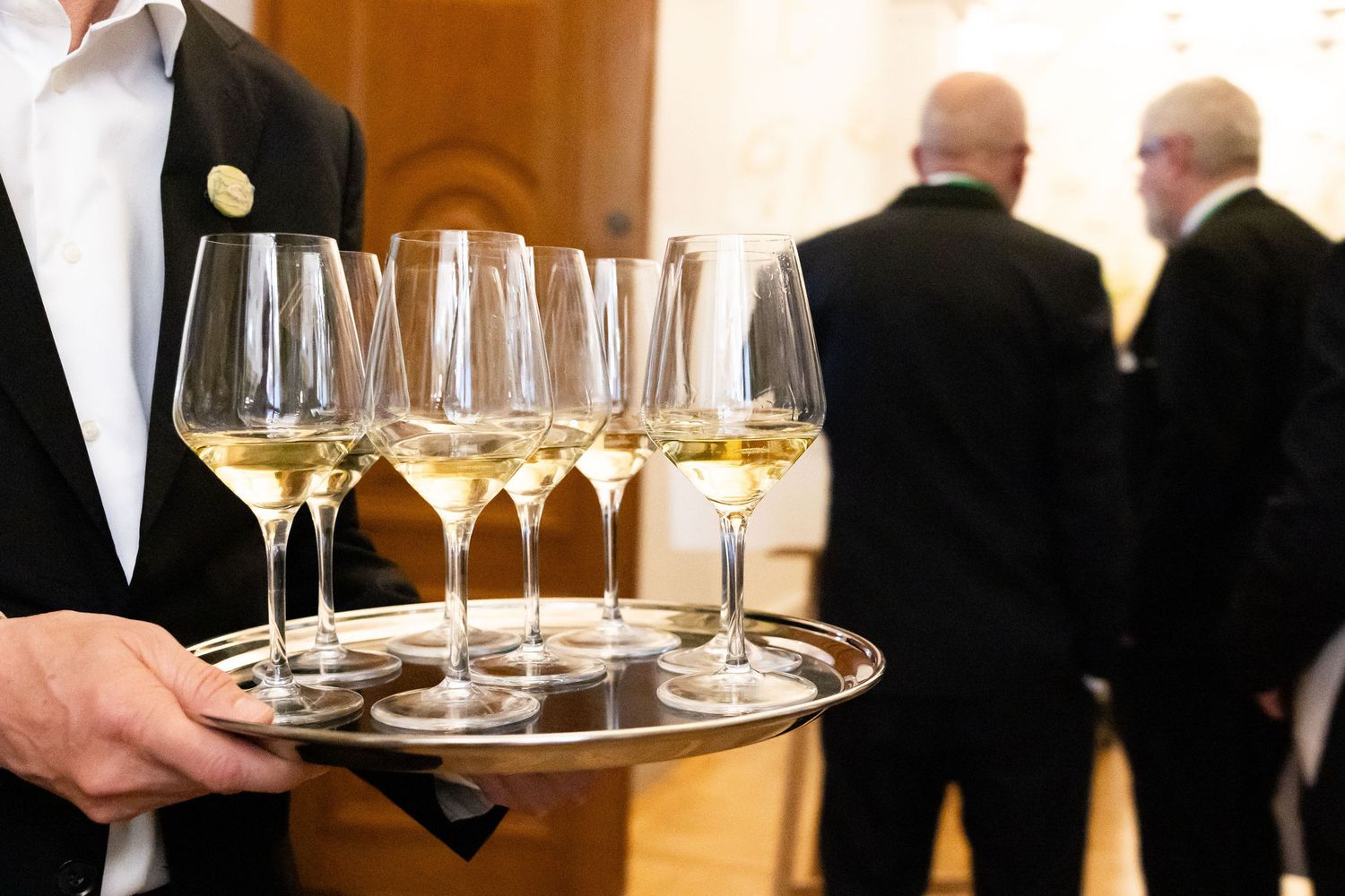 Waiter in formal attire carrying a silver tray of white wine glasses at an elegant event.