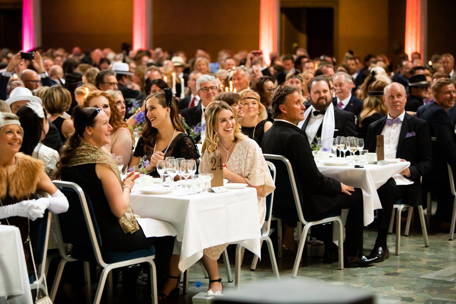 Elegantly dressed guests enjoying a formal gala dinner, seated at white-clothed tables in a grand ballroom.