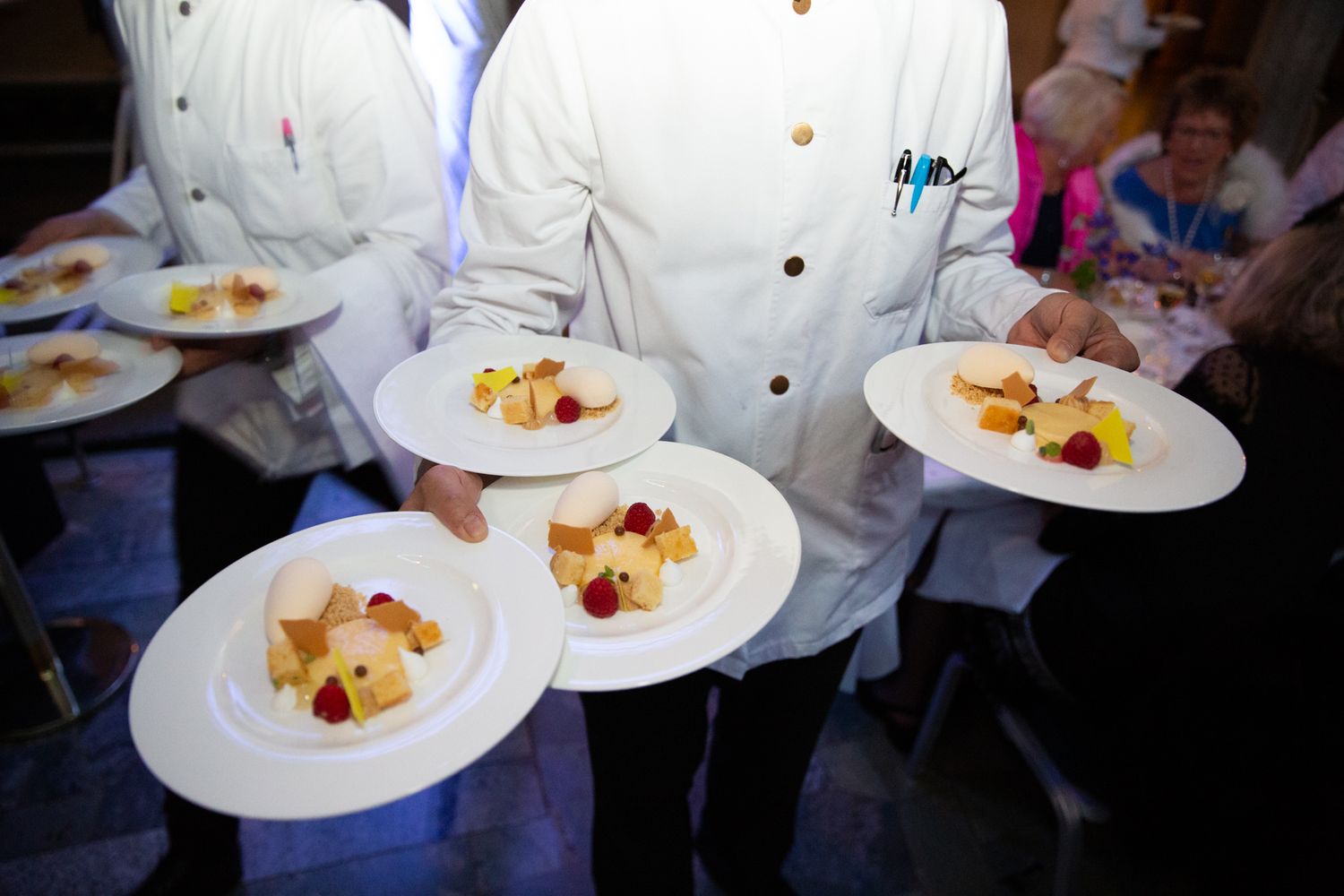 Chefs in white uniforms carrying elegant dessert plates with fresh fruit at a catered event.