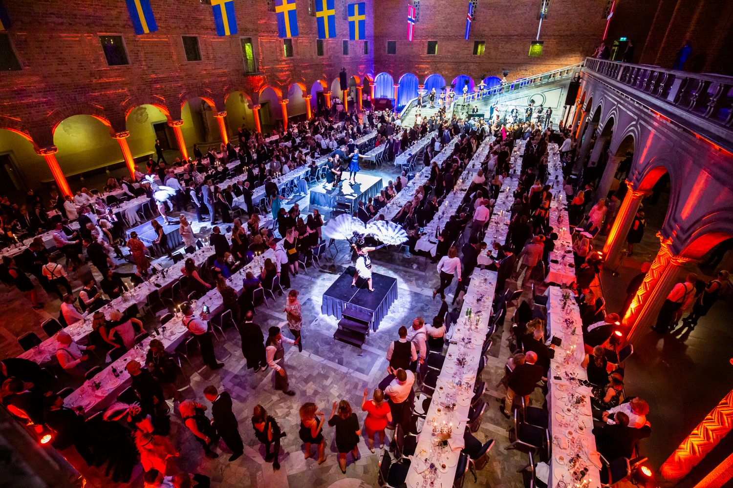 Aerial view of an elegant banquet hall with long dining tables, guests, and a glowing stage under Swedish flags.