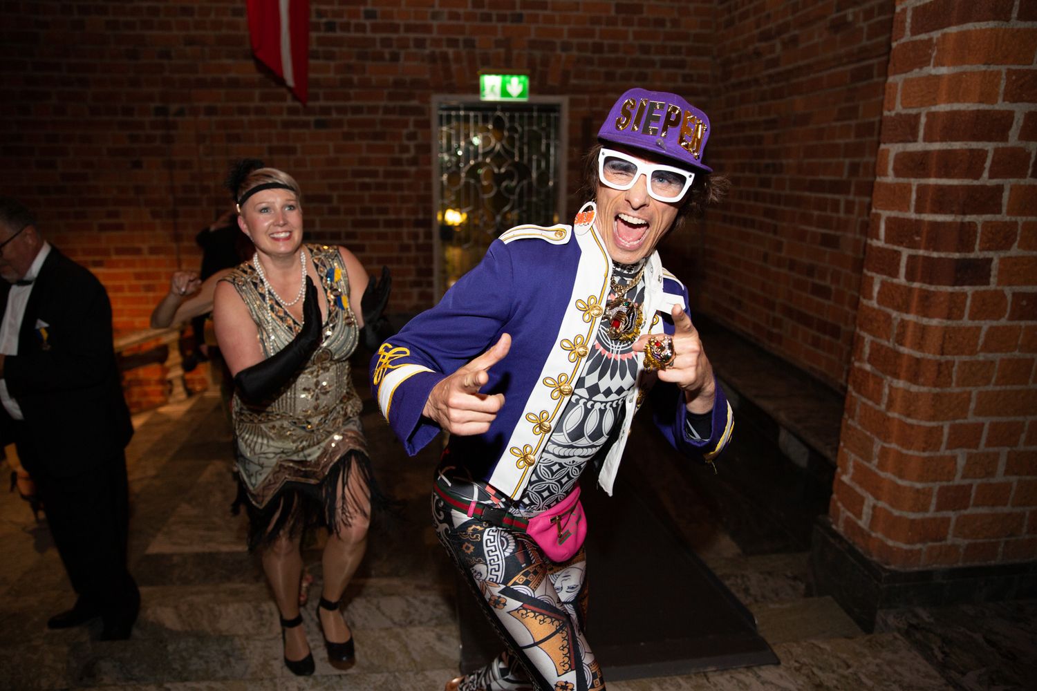 Two people in colorful costumes dancing at a festive party event in a brick venue.