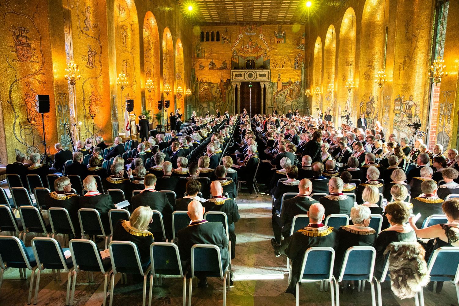 Elegant concert hall filled with audience seated in rows, featuring ornate golden walls and murals during a formal event.