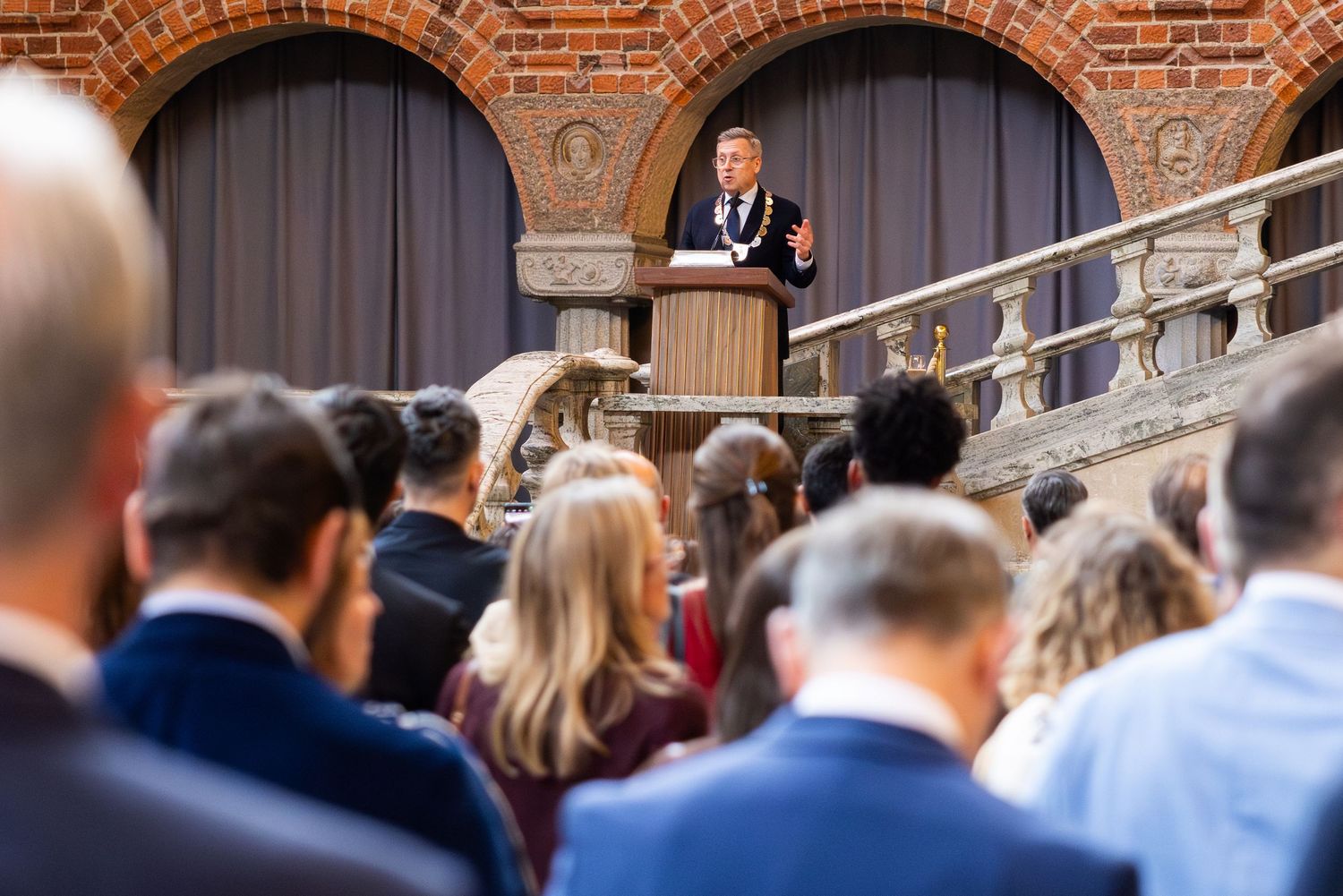A speaker addresses a large audience at an outdoor academic ceremony near a historic brick building with arched windows.