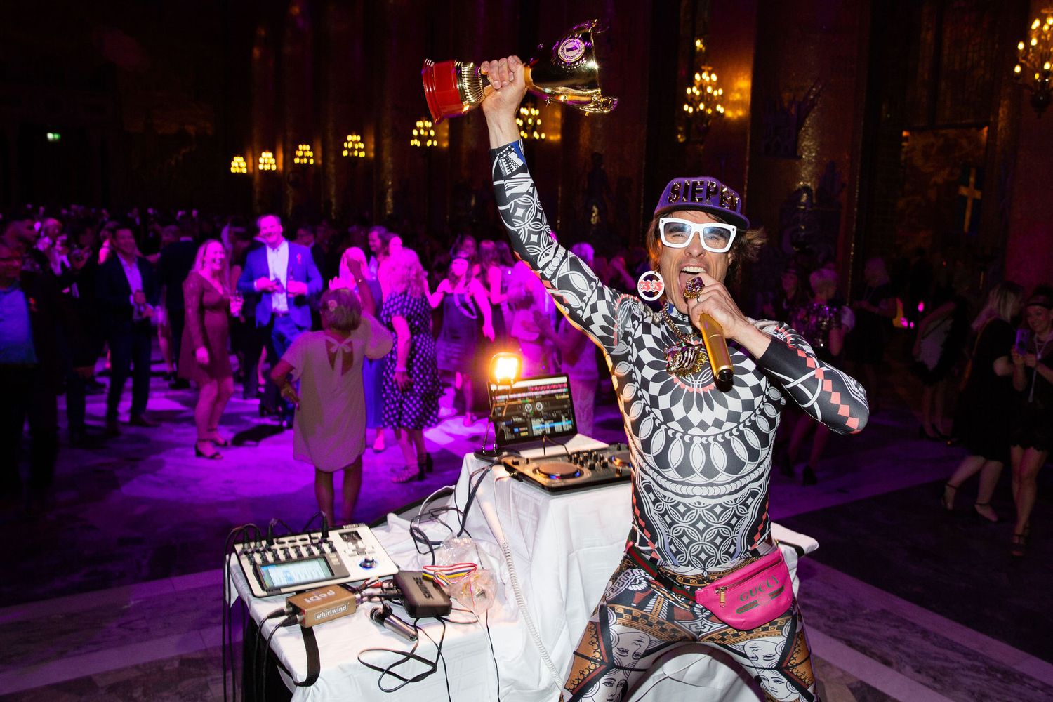 Energetic DJ in patterned outfit performs at a vibrant purple-lit party venue with crowd dancing in background.