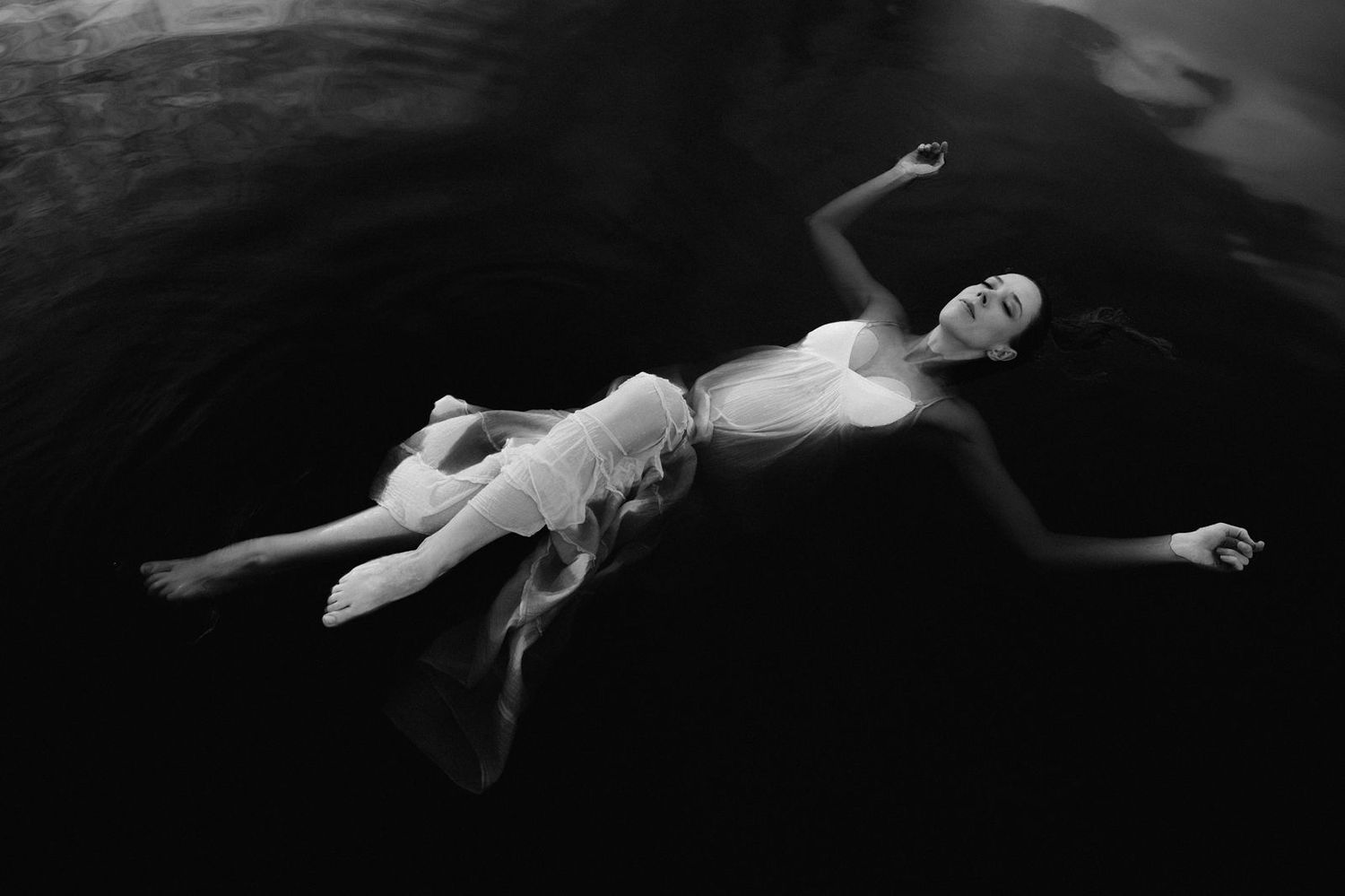 Black and white photo of a woman in a white dress floating gracefully on dark water.