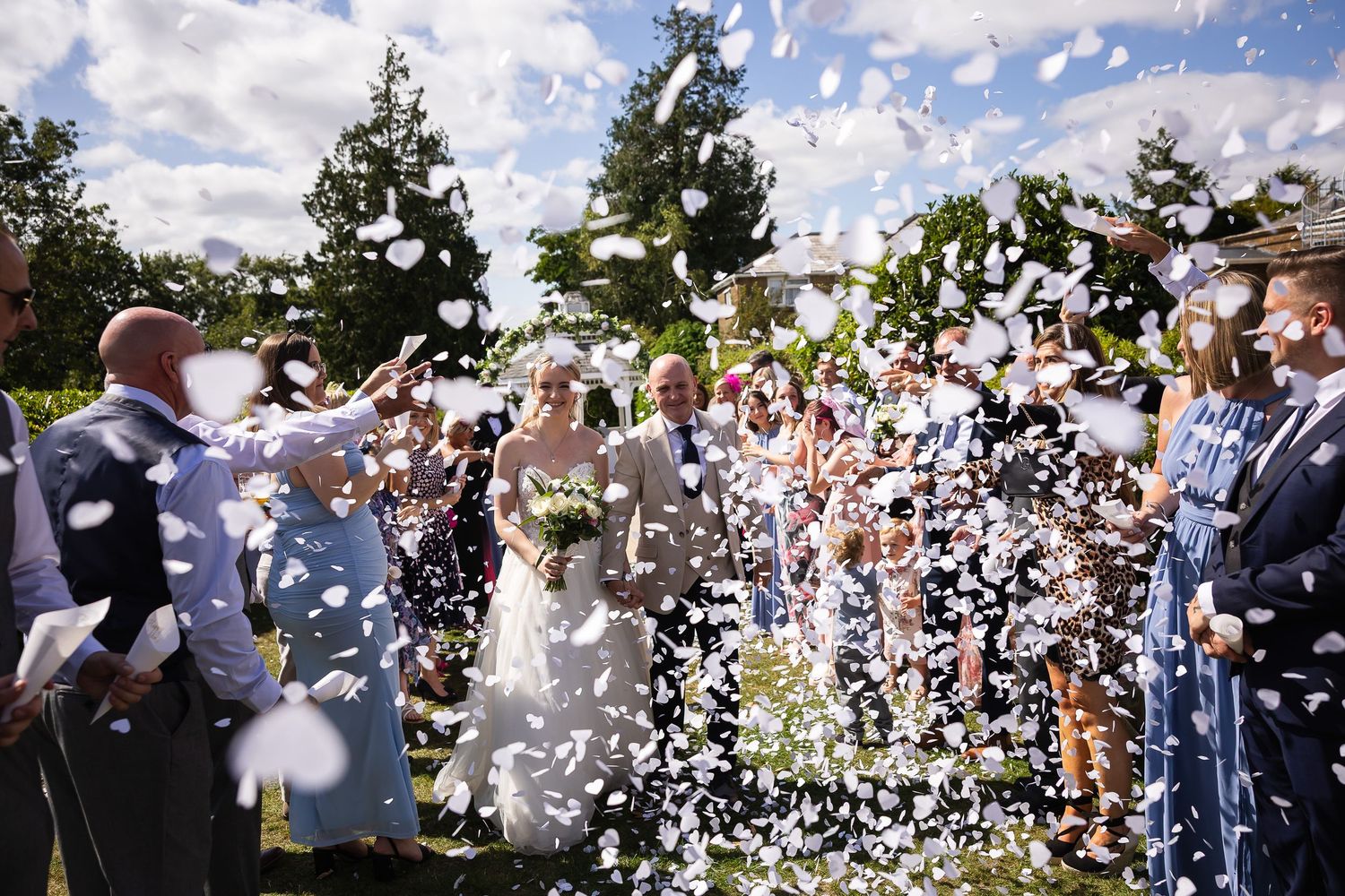 Bride and groom walk through confetti shower surrounded by cheering wedding guests outdoors on a sunny day.