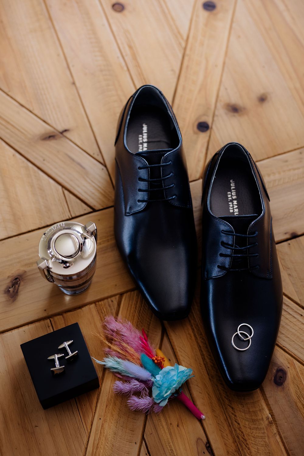 Black leather dress shoes with wedding rings, cufflinks, colorful boutonniere, and cologne on wooden floor.
