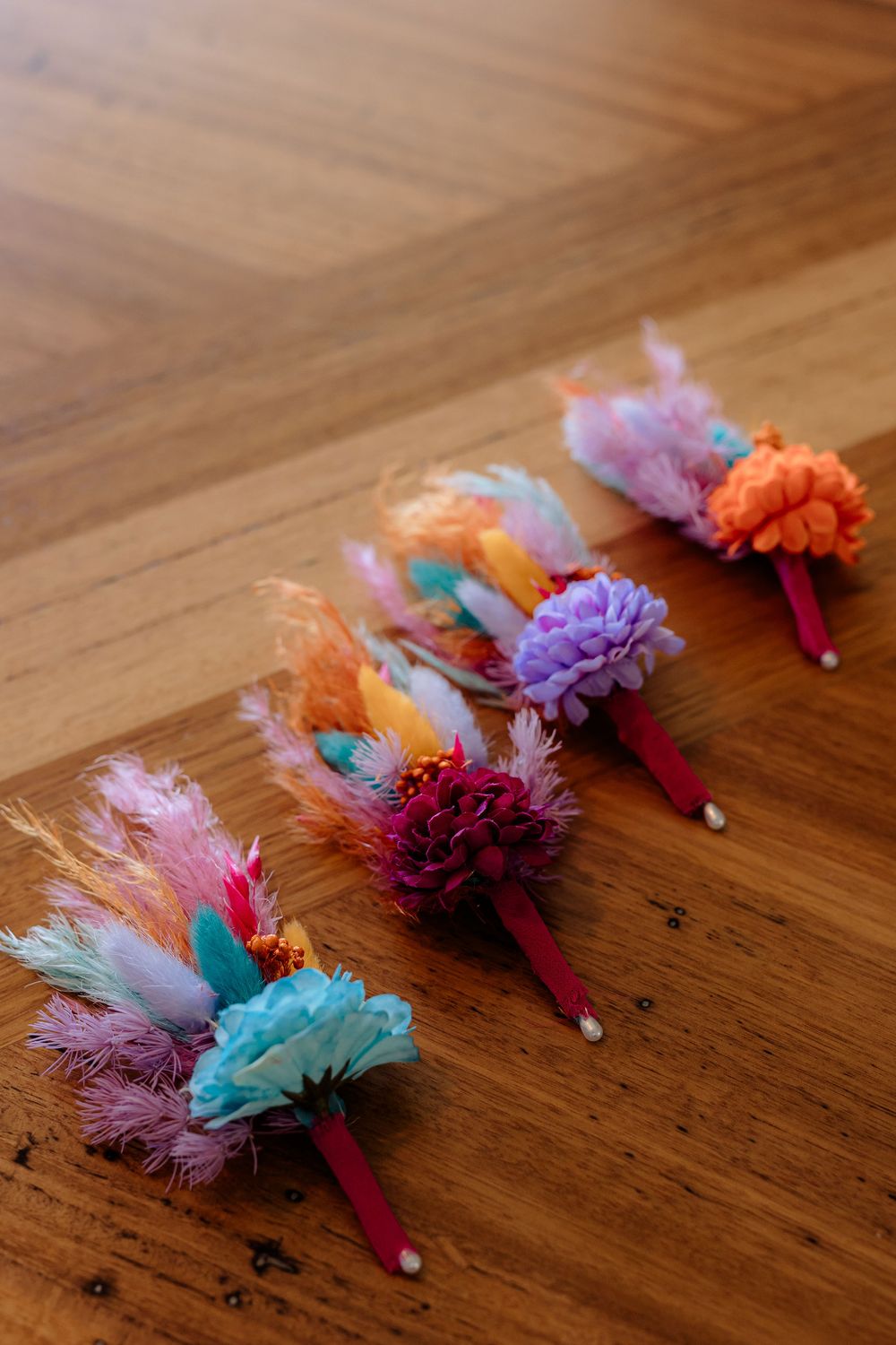 Colorful floral boutonnieres with feathers and mixed blooms arranged in a row on a wooden surface.
