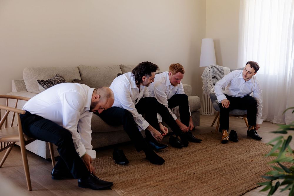 Four groomsmen in white shirts and black pants putting on dress shoes together in a bright modern living room.