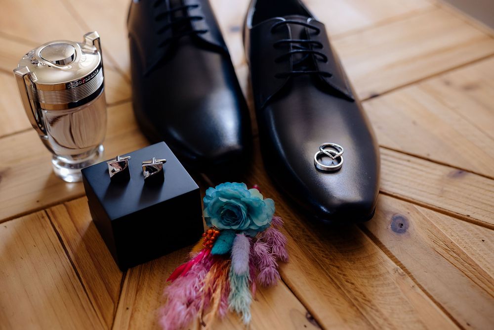 Black leather dress shoes with wedding rings, cufflinks, boutonniere, and cologne on wooden floor for groom wedding prep.