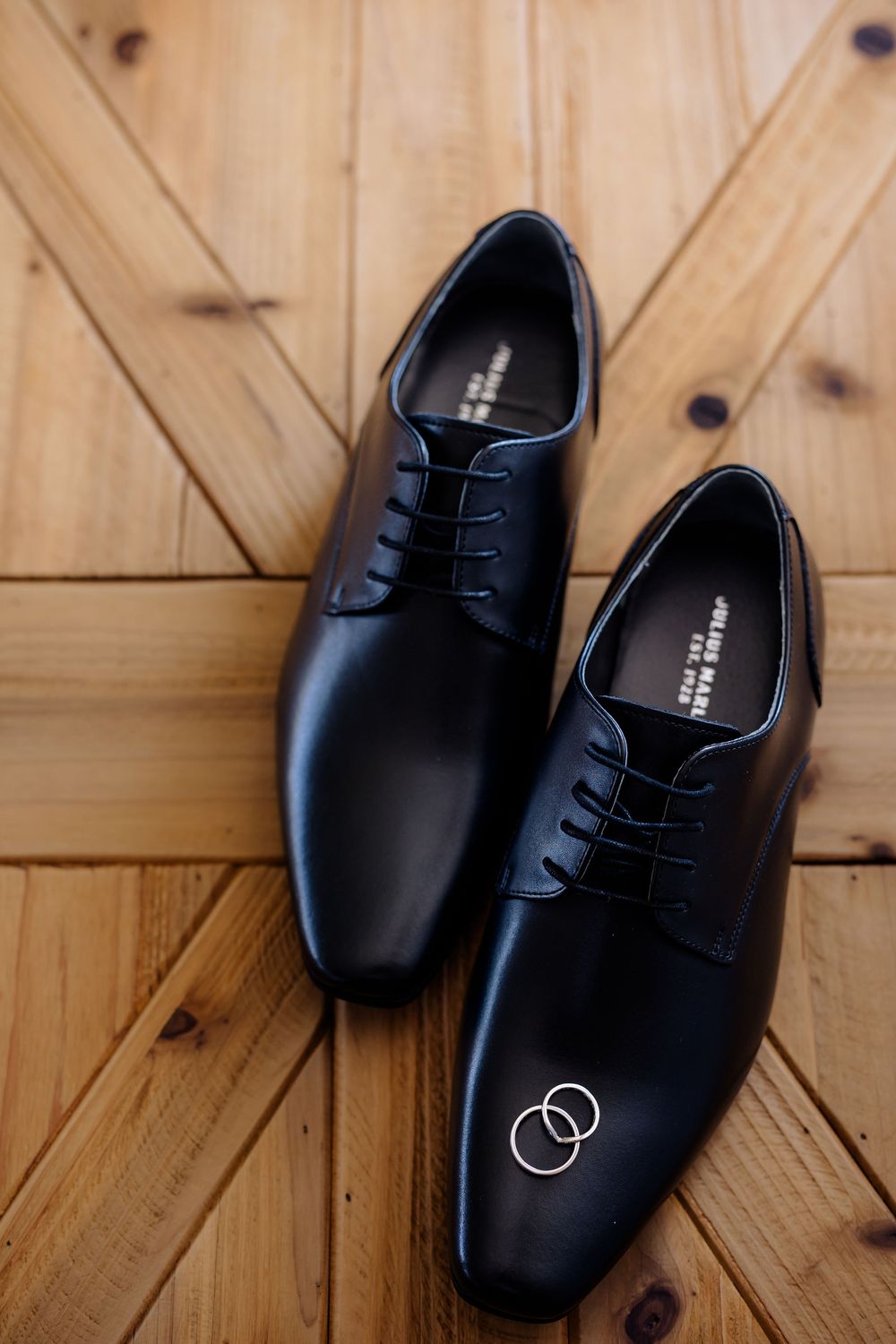Black leather dress shoes with wedding rings on wooden floor, perfect for groom wedding day accessories.