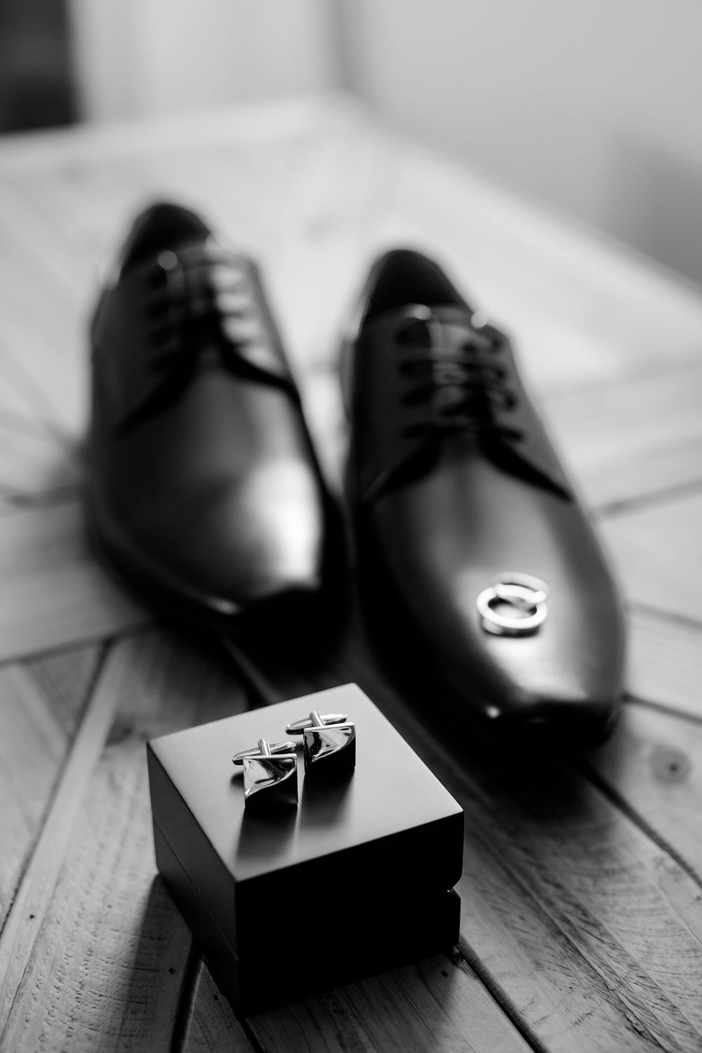 Black and white photo of polished dress shoes with cufflinks on a box and wedding rings, perfect for a groom's wedding day.