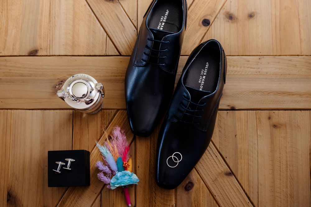 Black leather dress shoes displayed on wood floor with wedding rings, cufflinks, boutonniere, and cologne for groom.