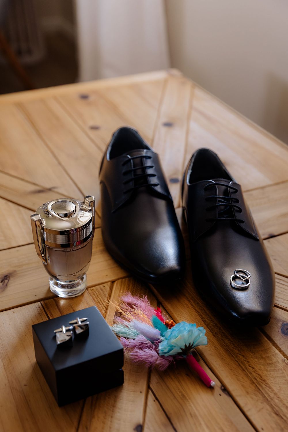 Black leather dress shoes, cologne, cufflinks, wedding rings, and a colorful boutonniere arranged on a wooden table.