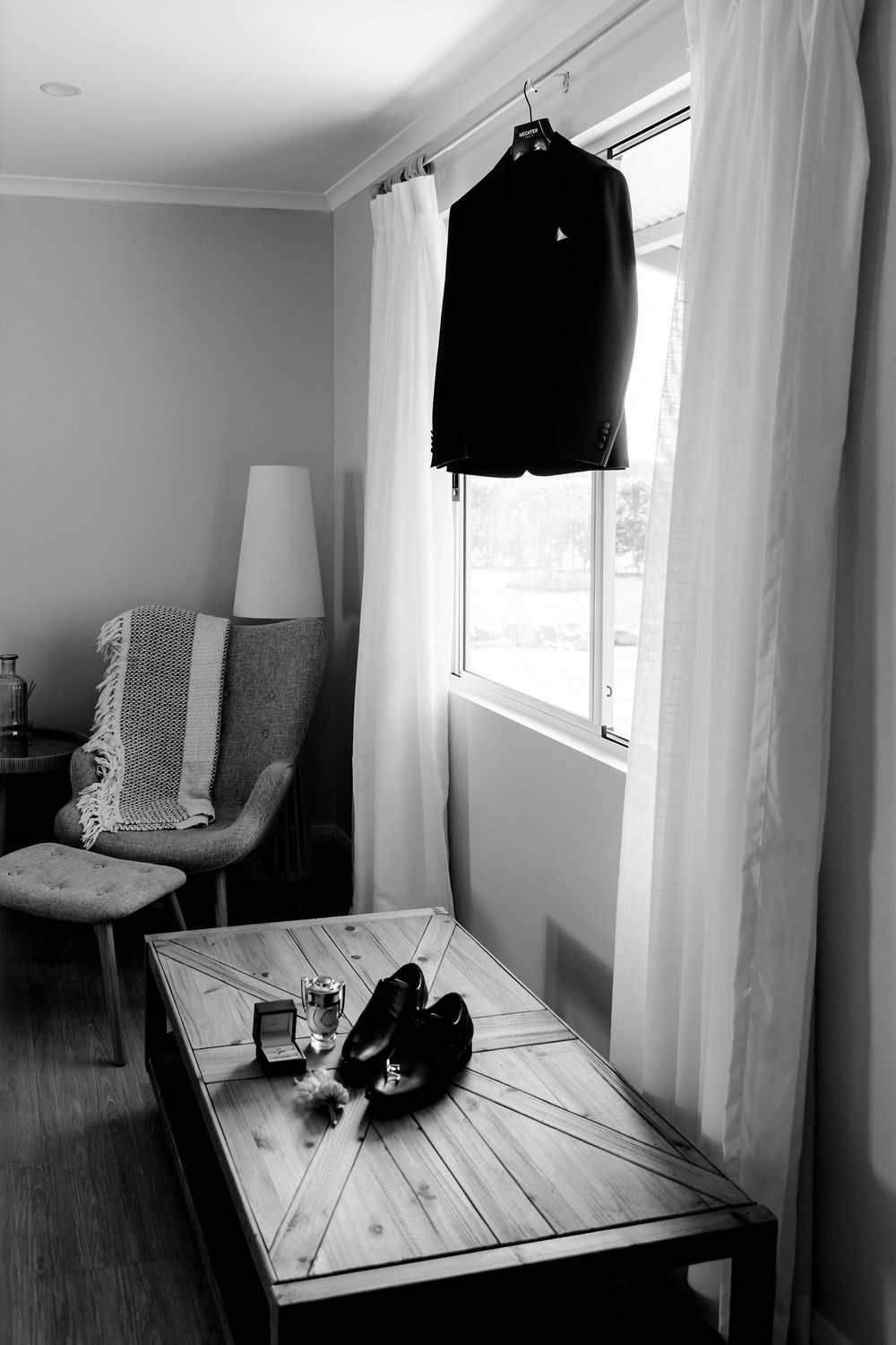 Black and white photo of a groom's suit jacket hanging by window with shoes and ring on wooden table.
