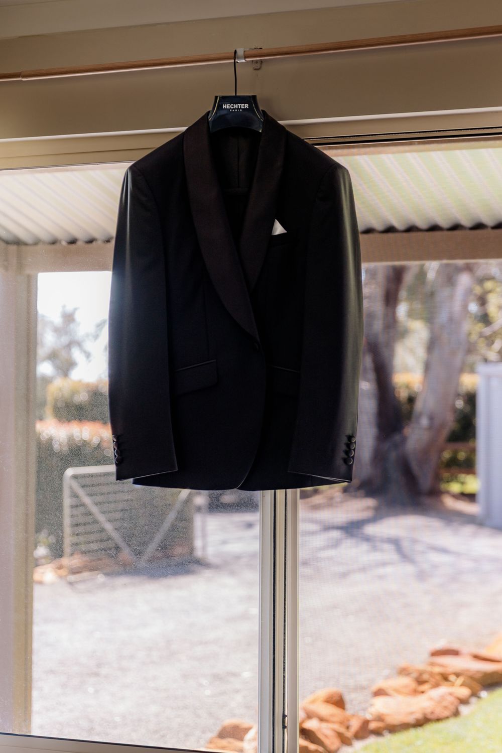 Black tuxedo jacket with white pocket square hanging on hanger near sunlit window with garden view.