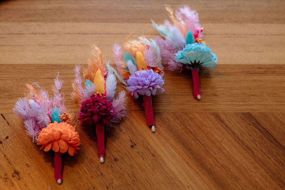 Four colorful floral boutonnieres with feathers and flowers on red stems arranged on a wooden surface.