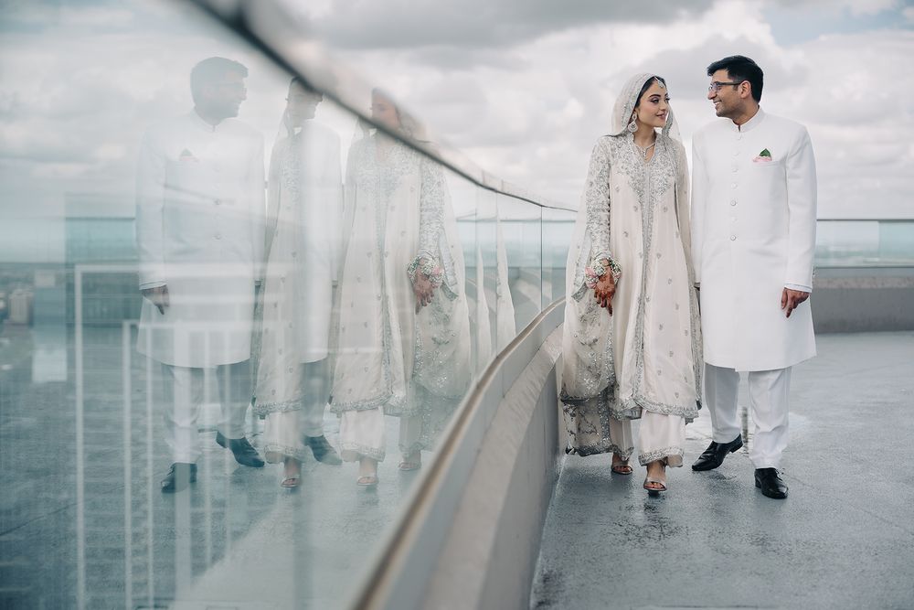 South Asian bride and groom in white traditional wedding attire standing on a rooftop with glass railings.