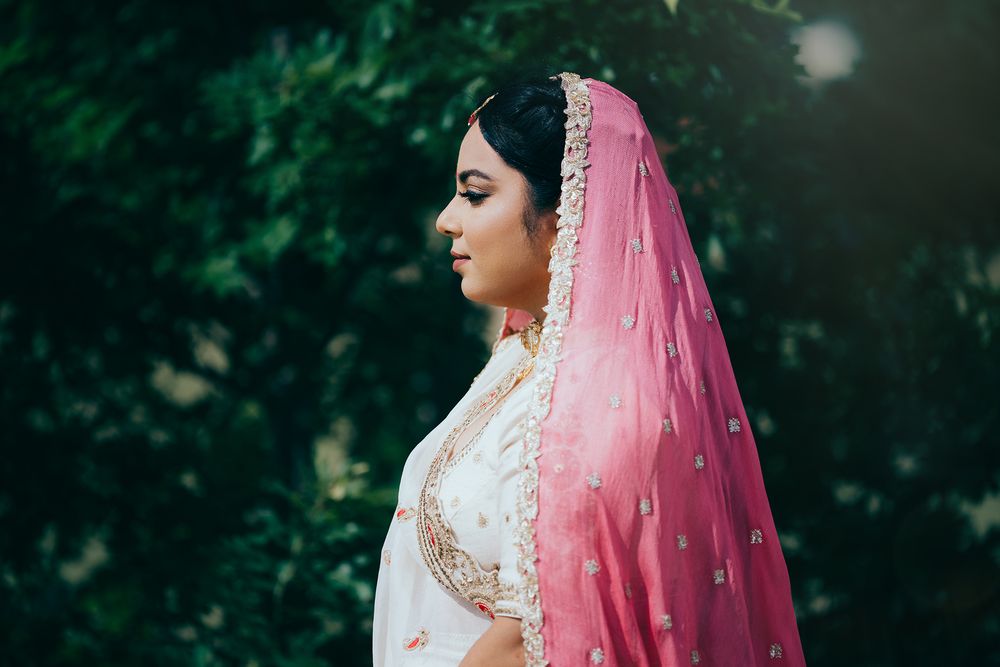 South Asian bride in white and gold lehenga with pink dupatta veil poses gracefully outdoors amid lush green foliage.