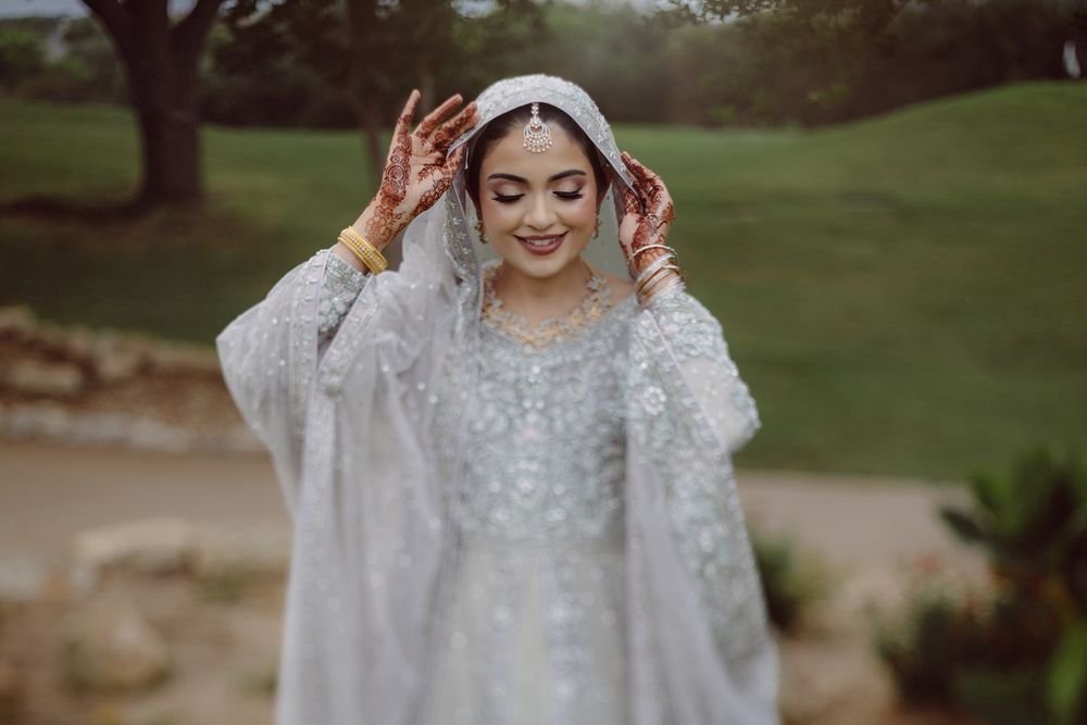South Asian bride in white embroidered lehenga adjusting her veil, adorned with mehndi and gold jewelry outdoors.