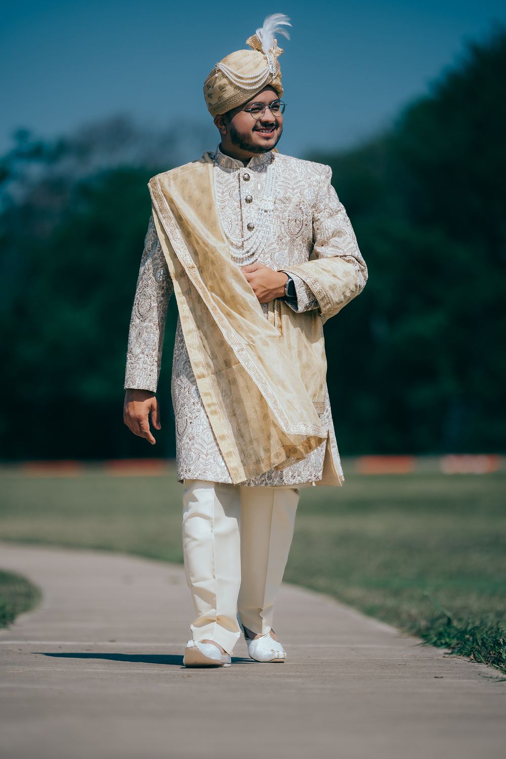 Indian groom in ornate cream sherwani and golden turban with feather walking outdoors on sunny day.