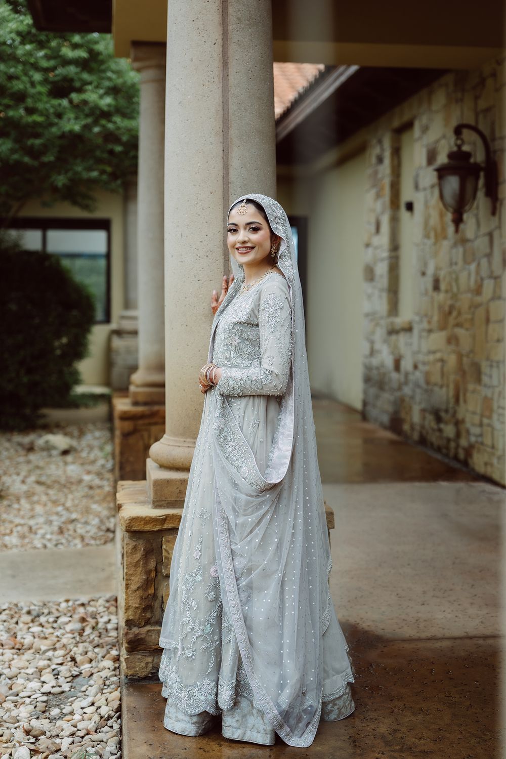 Bride in silver embroidered South Asian bridal gown with dupatta, posing by stone columns outdoors.
