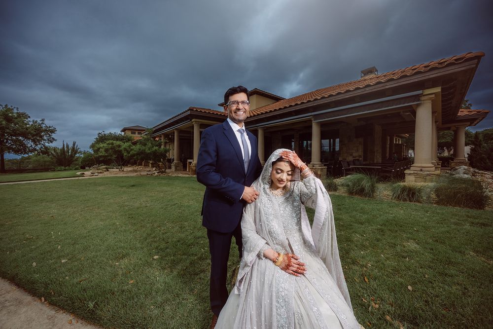 South Asian couple in wedding attire pose outdoors, groom in navy suit, bride in silver lehenga, with dramatic stormy sky.