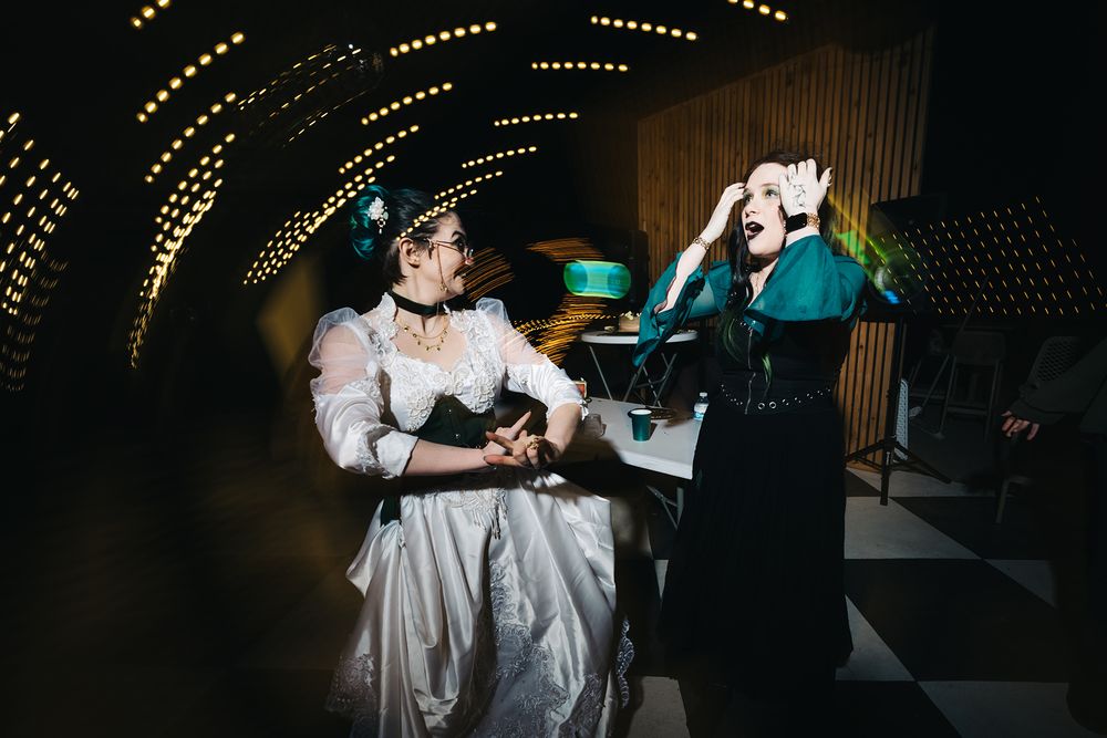 Two women in elegant costumes laugh and dance in a dramatic tunnel venue with glowing lights.