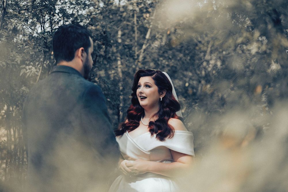 Bride in off-shoulder white gown smiles at groom amid golden foliage in romantic outdoor wedding portrait.