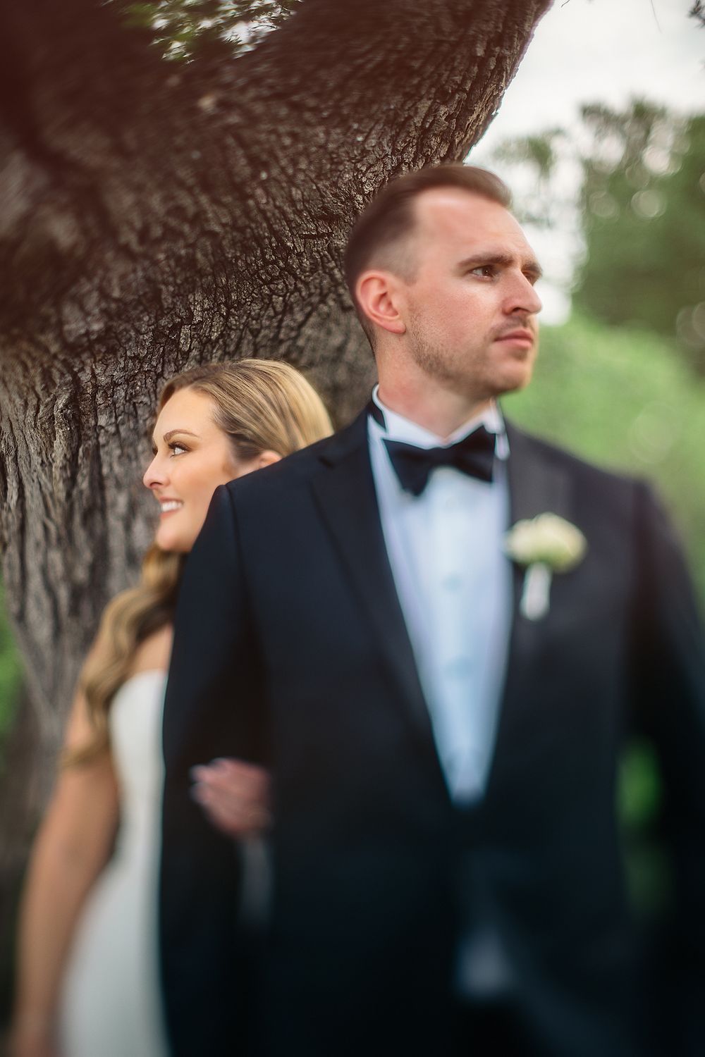 Bride and groom pose by a large tree, groom in black tuxedo with bow tie, bride in white gown smiling softly.