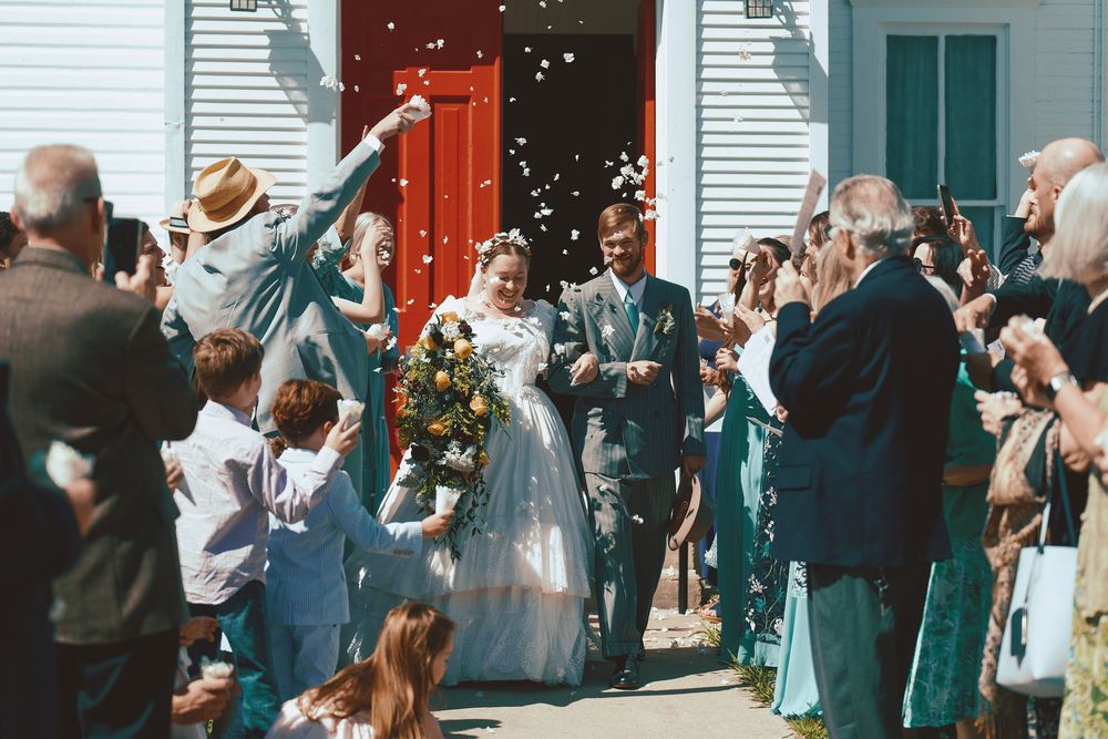 Joyful newlyweds exit church as guests toss rice in sunny outdoor wedding celebration.