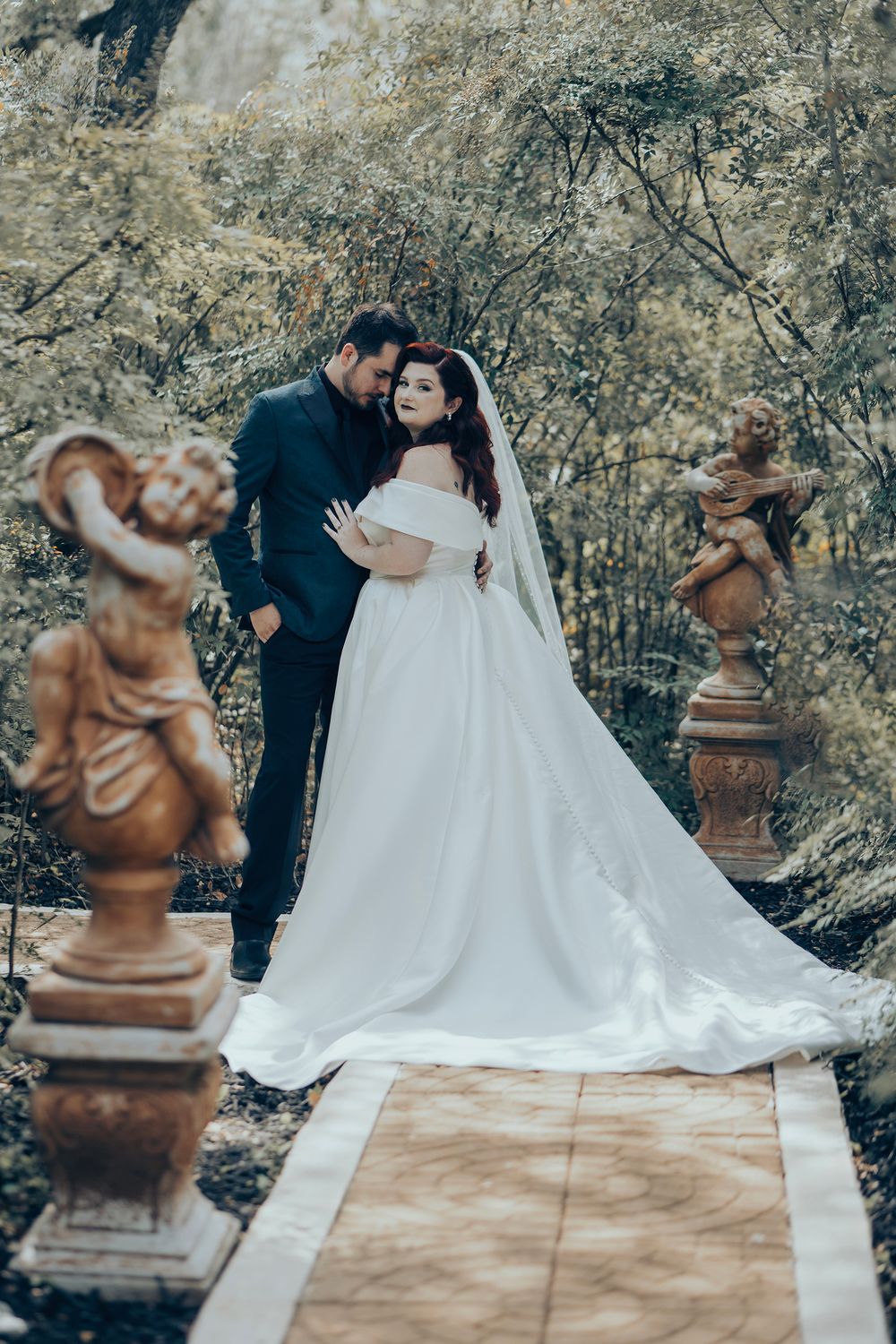 Bride and groom embrace in an enchanting garden with cherub statues, bride wearing an elegant white ball gown with long train.