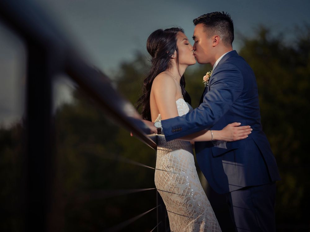 A couple shares a romantic kiss at their wedding, with the groom in a blue suit and bride in a white gown outdoors.