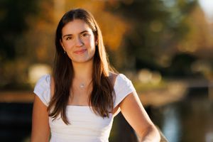 Young woman with long brown hair wearing a white top, smiling outdoors with autumn foliage background.