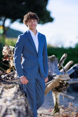 Young man in blue pinstripe suit smiling outdoors near driftwood logs with trees in background.