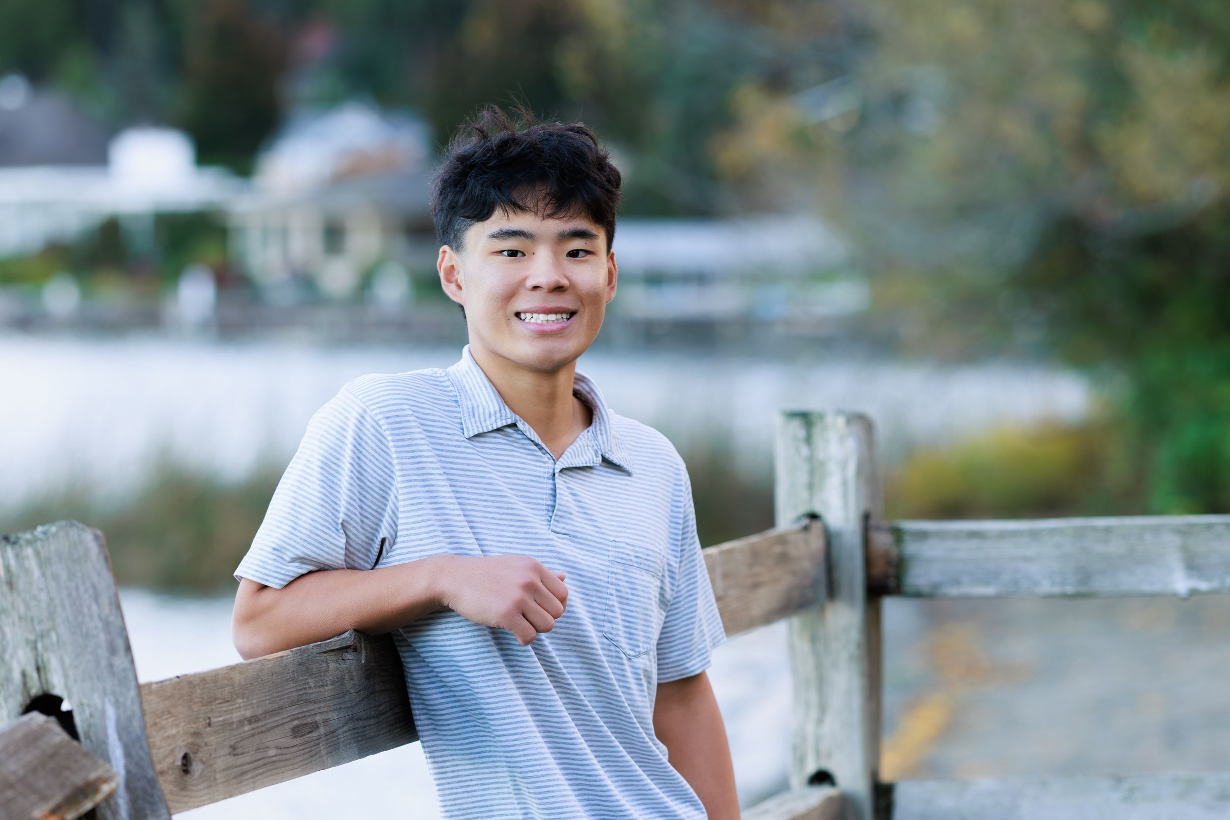 Smiling young Asian man leaning on wooden fence outdoors near water, wearing gray polo shirt.