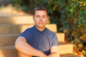 Young man with crossed arms sitting on stone steps outdoors, wearing a blue patterned shirt in warm sunlight.